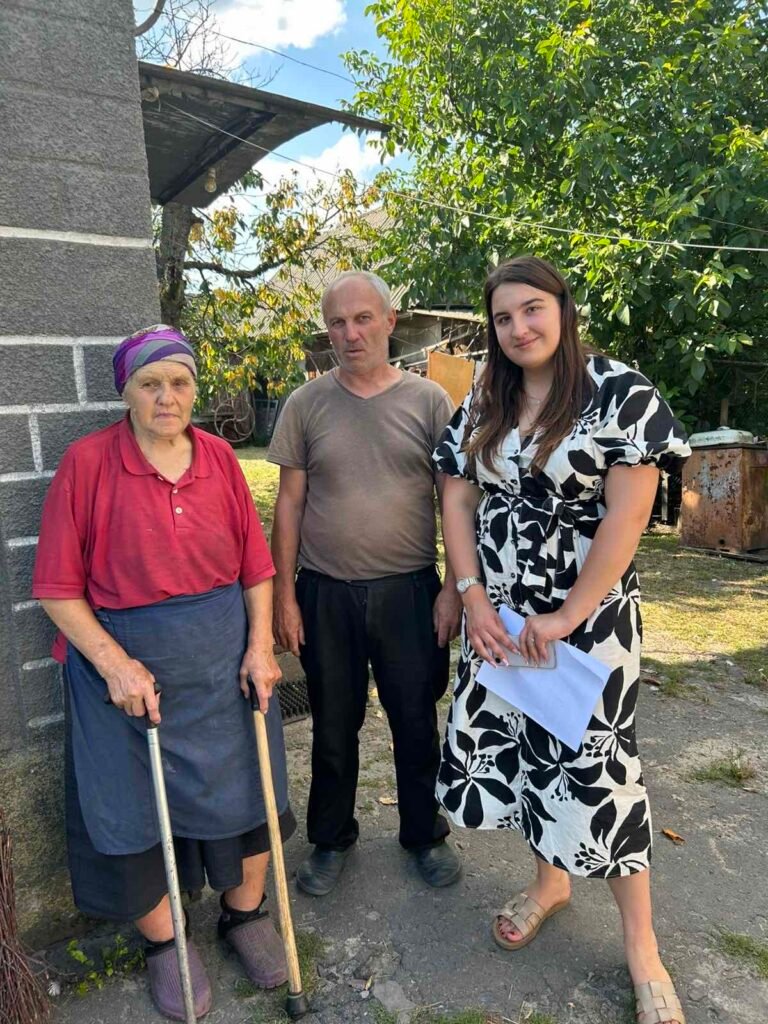 Троє людей стоять на подвірʼї поруч із кам’яною стіною будинку. Ліворуч – літня жінка у червоній сорочці, темній спідниці, з деревʼяними палицями для опори, на голові у неї хустка. Посередині стоїть сивий чоловік у сірій футболці та чорних штанах. Праворуч – молода жінка у білій сукні з чорними квітковими візерунками, у руках тримає аркуш паперу. Всі троє дивляться в камеру. Позаду видно зелень дерев і ділянку двору.