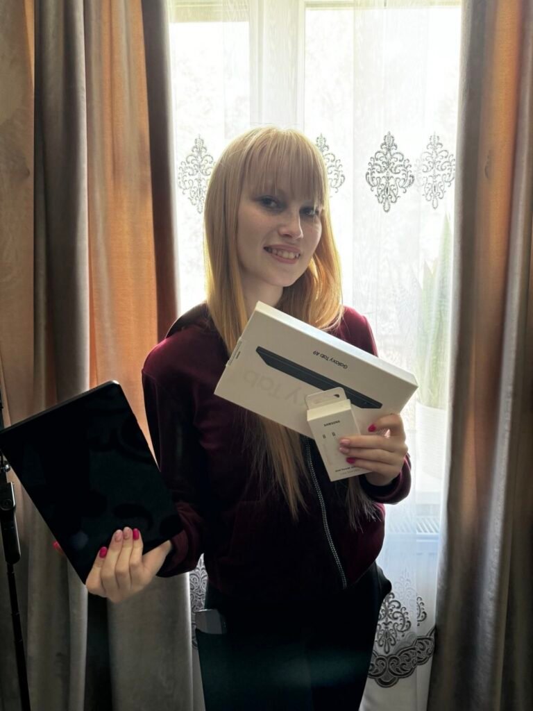 На фото Надія - дівчина з довгим світлим волоссям стоїть біля вікна з візерунчастими фіранками. Вона тримає в руках кілька речей: у правій руці коробку з написом 