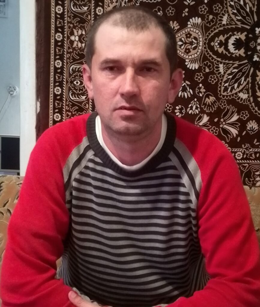 На фото: Олександр, Опішнянська громада. Чоловік сидить перед камерою на фоні килима з візерунком. Він має коротко стрижене волосся і вдягнений у светр з червоними рукавами та чорним корпусом у білі смужки. Його руки схрещені попереду себе. 