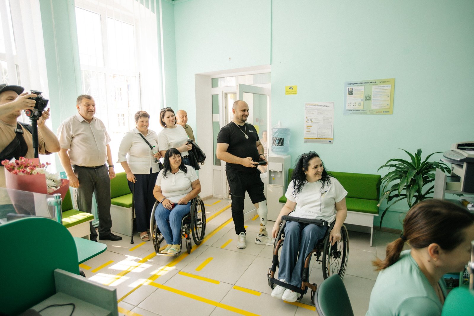 Учасники візиту перебувають у приміщенні установи у Сторожинецькій громаді. У кімнаті видно людей з інвалідністю, зокрема двох жінок на кріслах колісних, та інших учасників групи. Вони оглядають простір і спілкуються з працівниками установи. На підлозі нанесені жовті навігаційні лінії, на стіні розміщений інформаційний стенд. Атмосфера фото — робоча й доброзичлива, з акцентом на перевірку доступності послуг і приміщення.